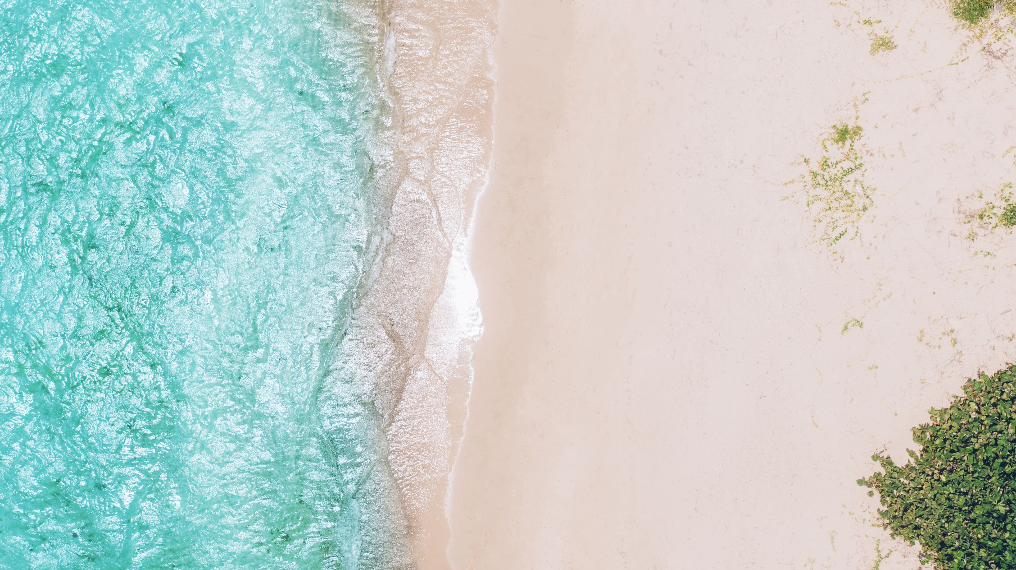 An aerial view of a beach with clear turquoise water, a sandy shore, and some greenery on the right side.