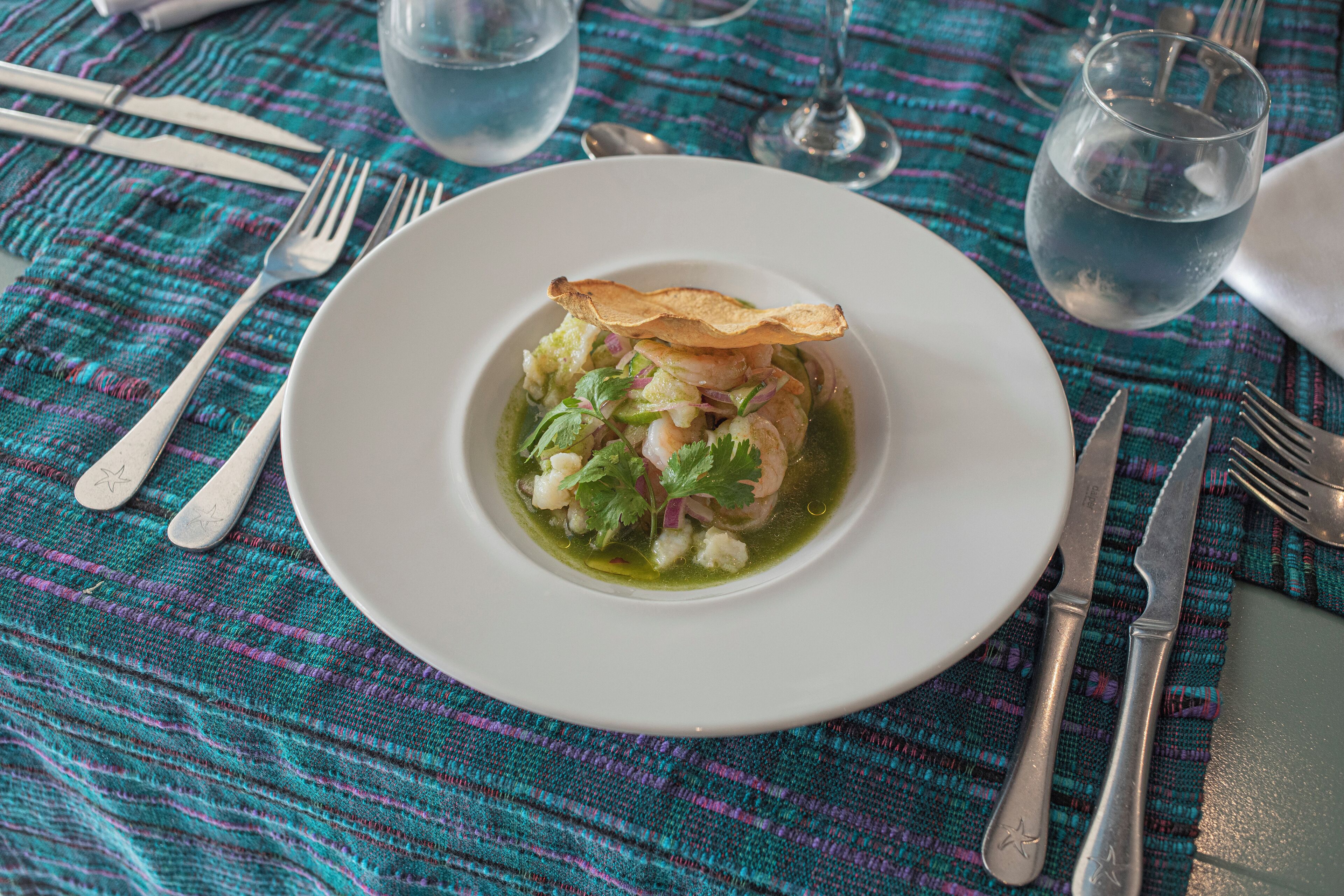 Une assiette blanche contenant un plat de fruits de mer, des couverts en argent et un verre d’eau, le tout posé sur une nappe à motifs tissés.