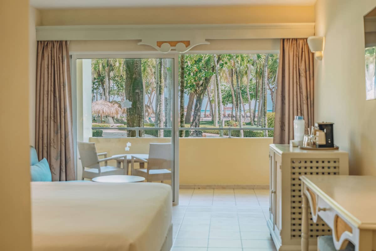 A hotel room with a bed, a small table, two chairs, a desk, and a window with a view of palm trees and a pool.