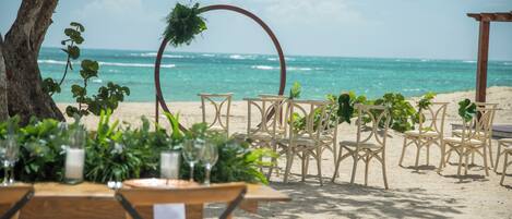 A beachside wedding setup with a wooden table and chairs, surrounded by greenery and a wooden arch.
