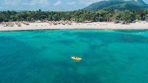 A beach with clear turquoise water, a person on a yellow raft, and a line of palm trees and beach huts in the background.
