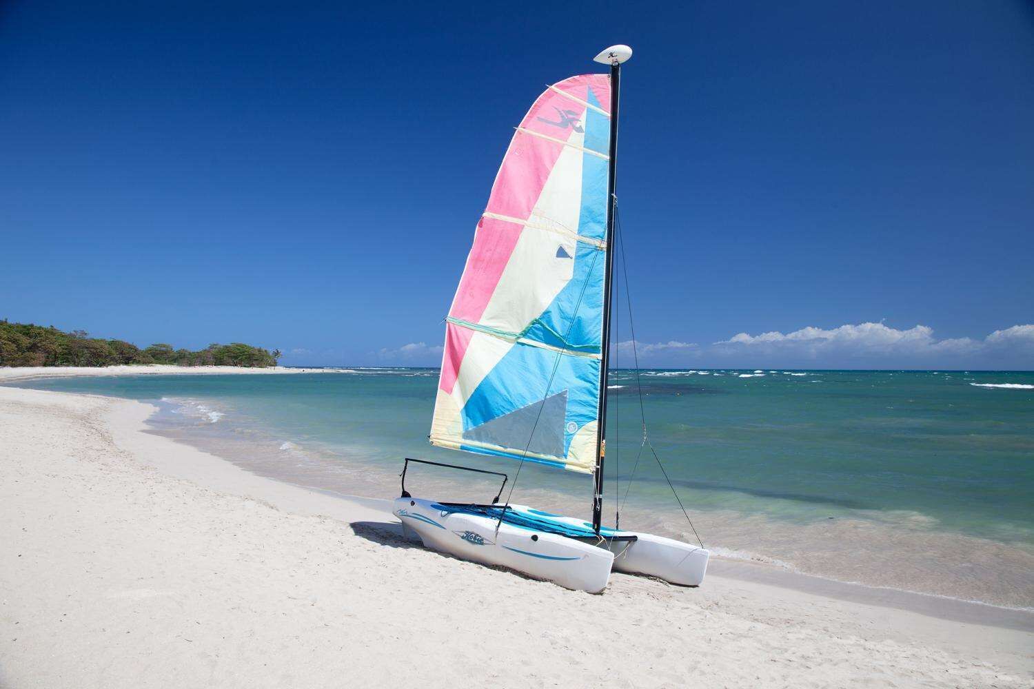 A sailboat with a colorful sail on a sandy beach with clear blue water and a distant shoreline.