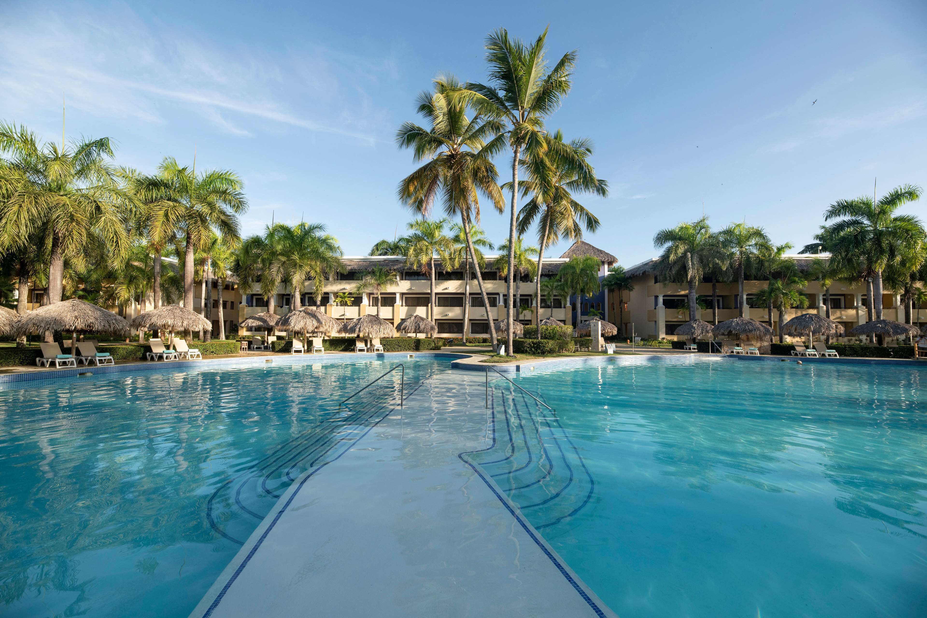 Ein großer Swimmingpool mit Liegestühlen und Palmen, umgeben von Resortgebäuden mit Strohdächern.