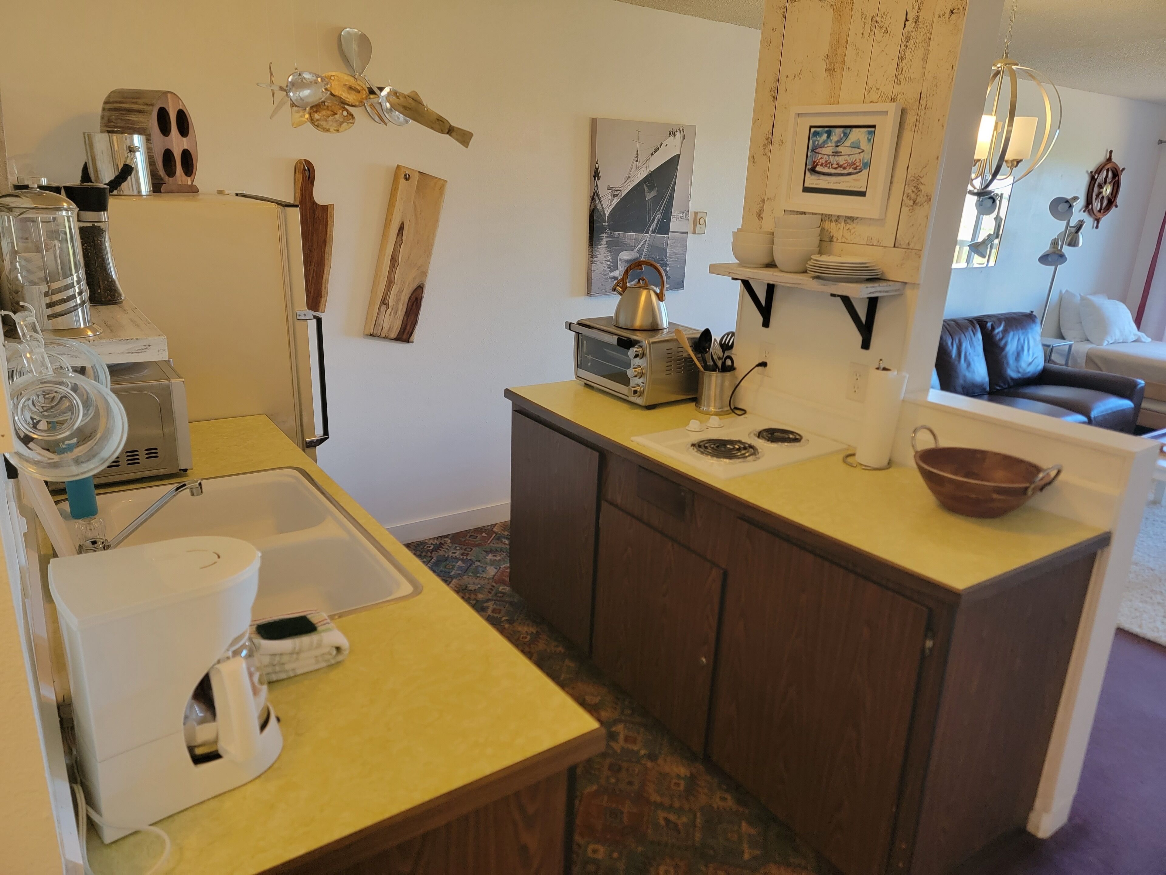 A compact kitchen with a sink, coffee maker, toaster, and a small stove. There are wooden cabinets and a window with a view of the outside.