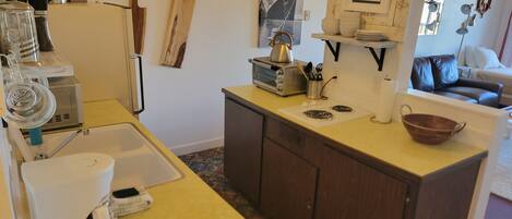 A compact kitchen with a sink, coffee maker, toaster, and a small stove. There are wooden cabinets and a window with a view of the outside.