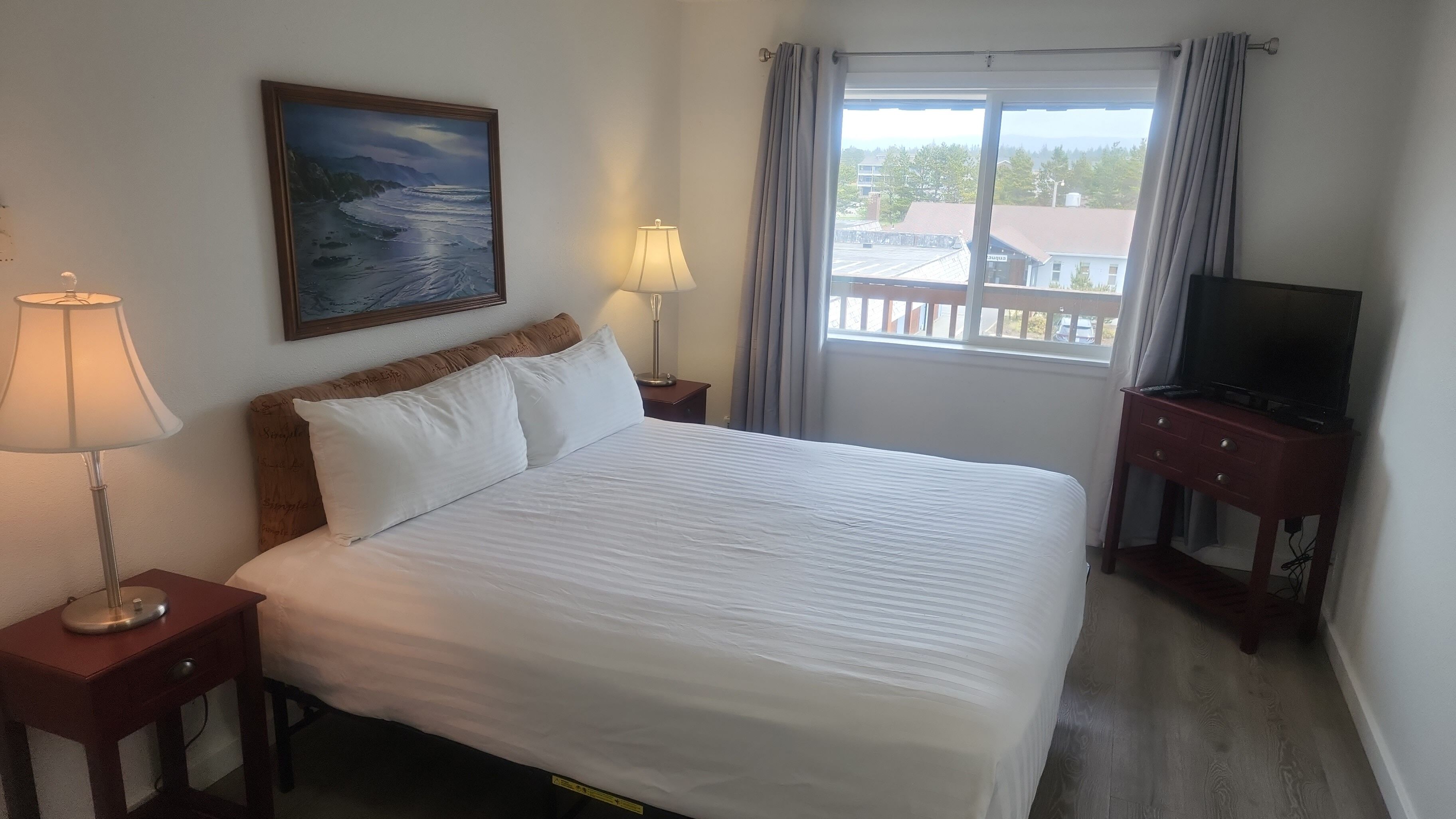 Habitación de hotel con cama, dos lámparas de noche, un cuadro, un televisor sobre un mueble y una ventana con cortinas.