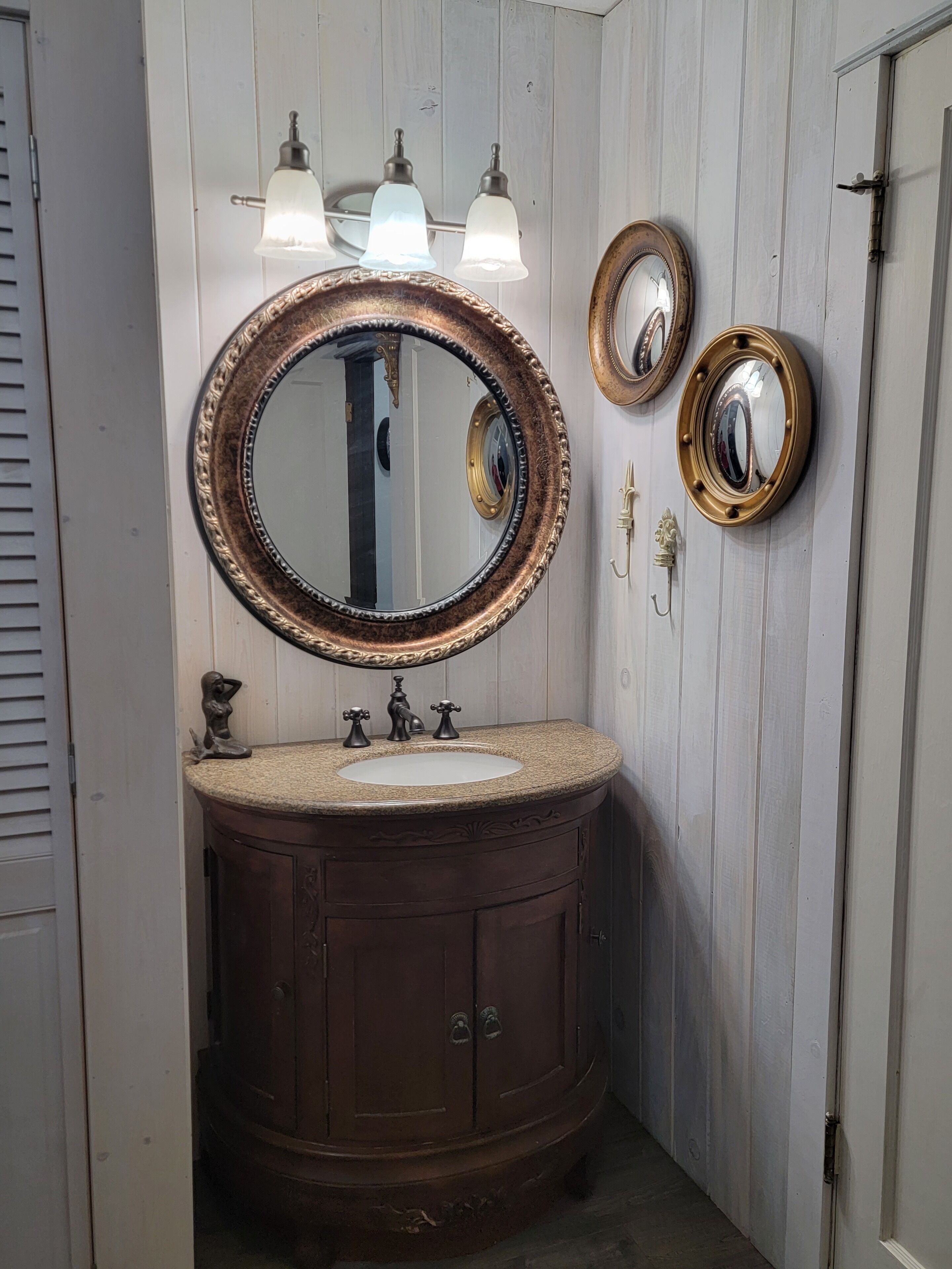 A bathroom with a dark wood vanity, a round mirror with a gold frame, and three wall-mounted lights.