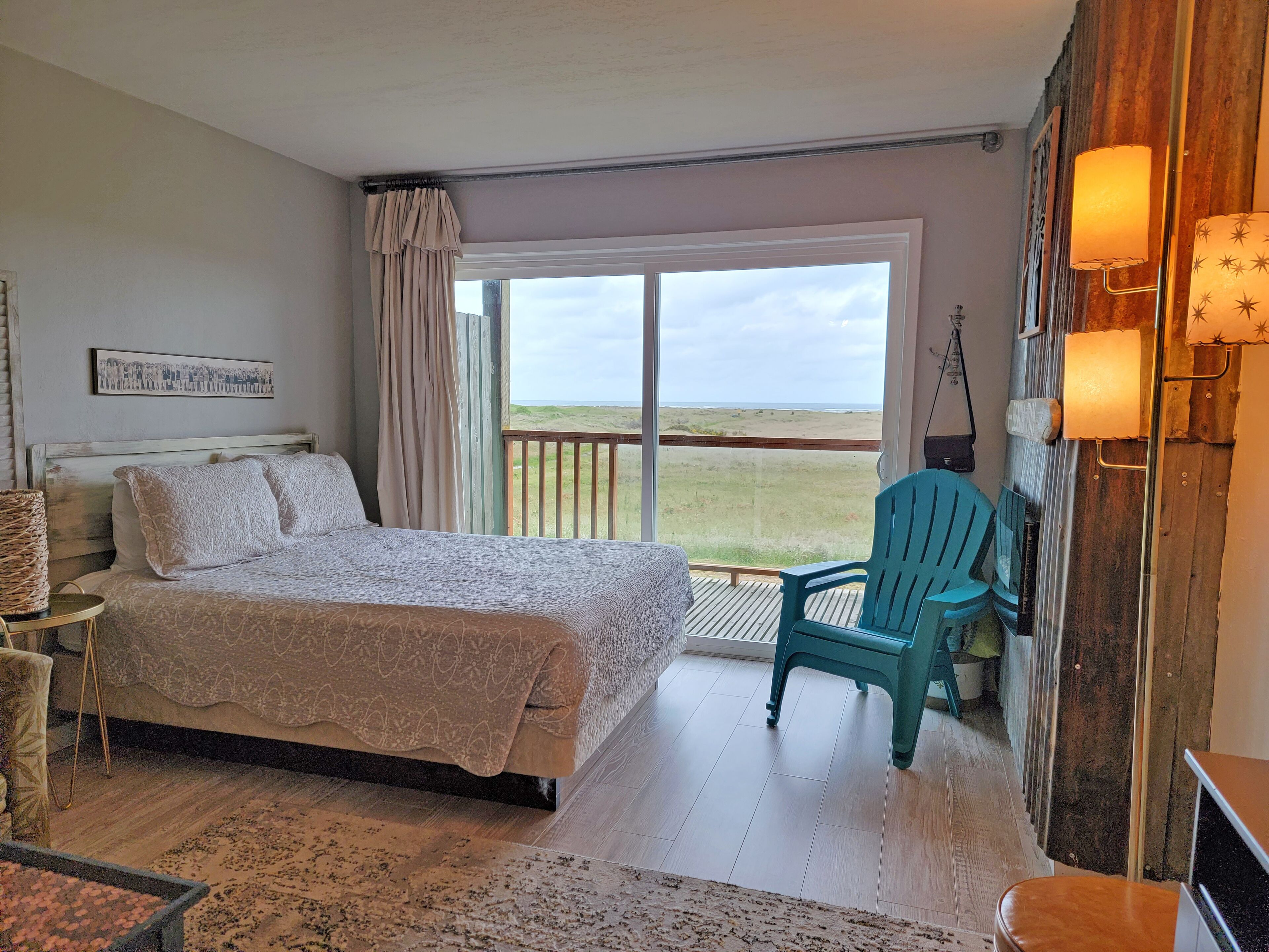 Un dormitorio con cama, una silla, un ventanal con vista a la playa y un balcón con barandilla.