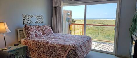 A bedroom with a bed, a nightstand, a lamp, a window with a view of the beach, and a sign that reads "BEACH LIVING".
