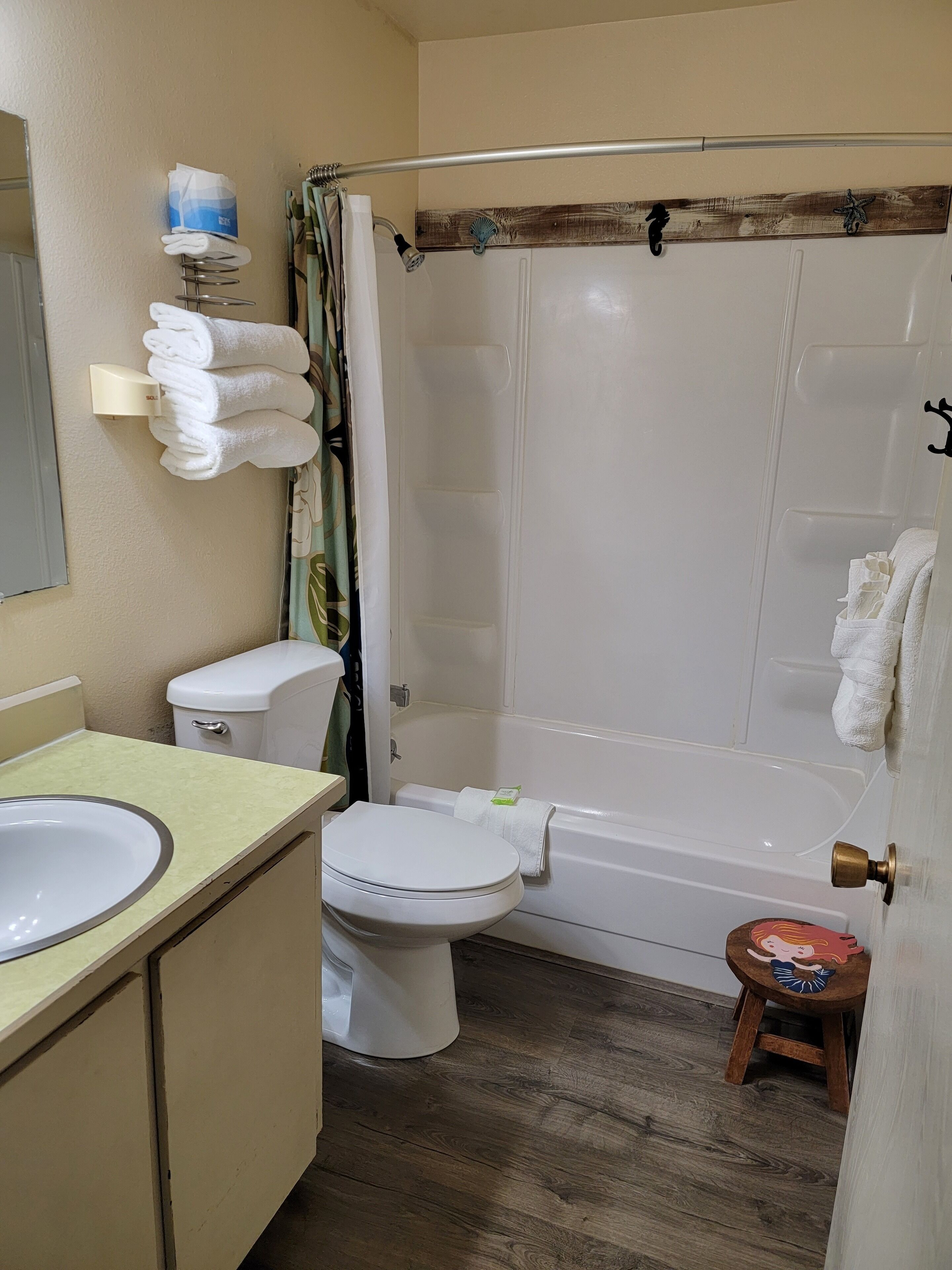 A bathroom with a toilet, sink, and bathtub. There is a towel rack with folded towels, a shower curtain, and a small stool with a decorative item.