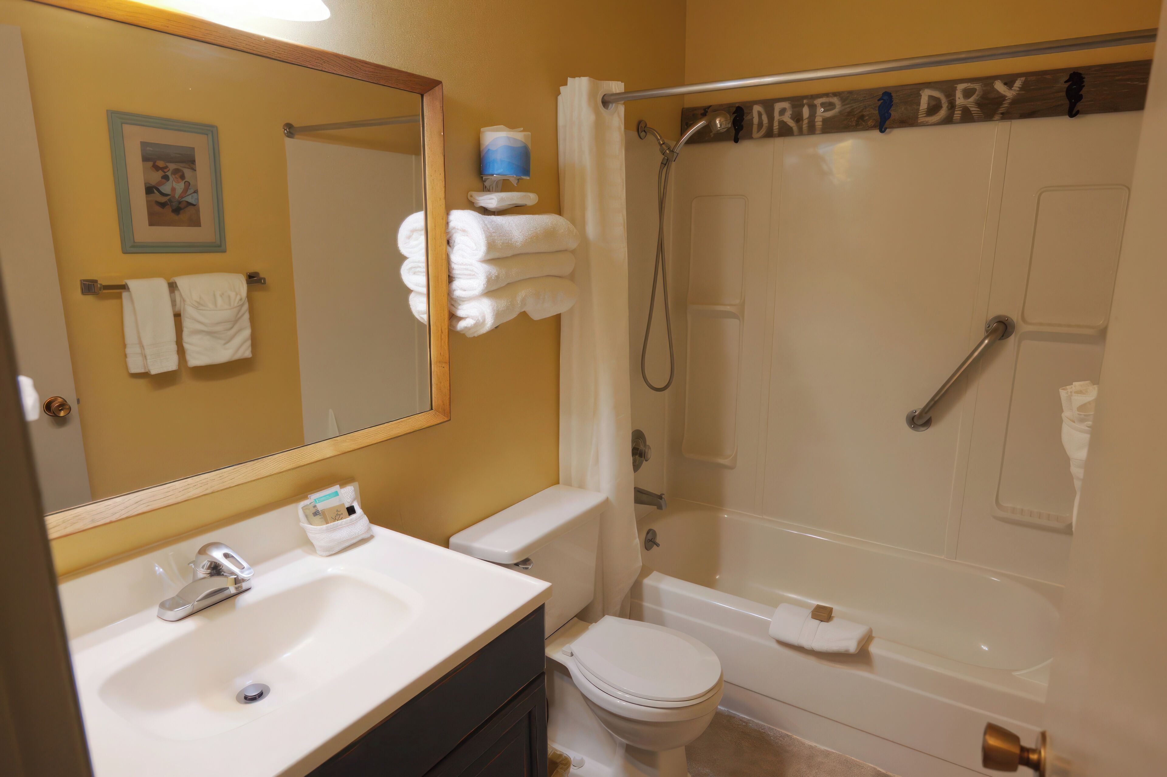 A bathroom with a sink, toilet, and bathtub. There is a towel rack with towels, a mirror, and a sign above the bathtub that says "DRIP DRY".