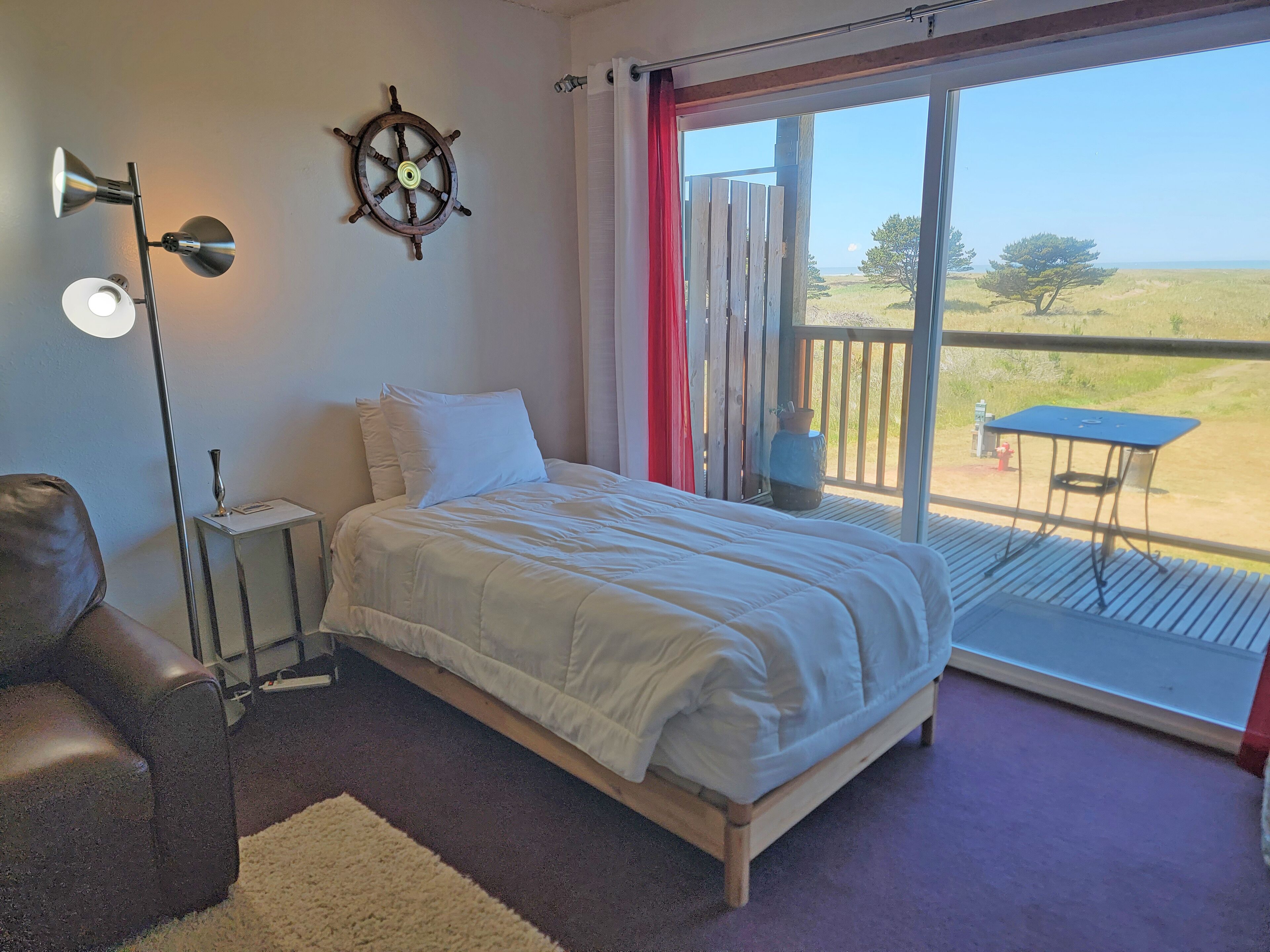 Une chambre à coucher comprenant un lit, une table de chevet, une lampe, un élément de décoration représentant un gouvernail de bateau, et une vue sur l’extérieur grâce à une porte coulissante.