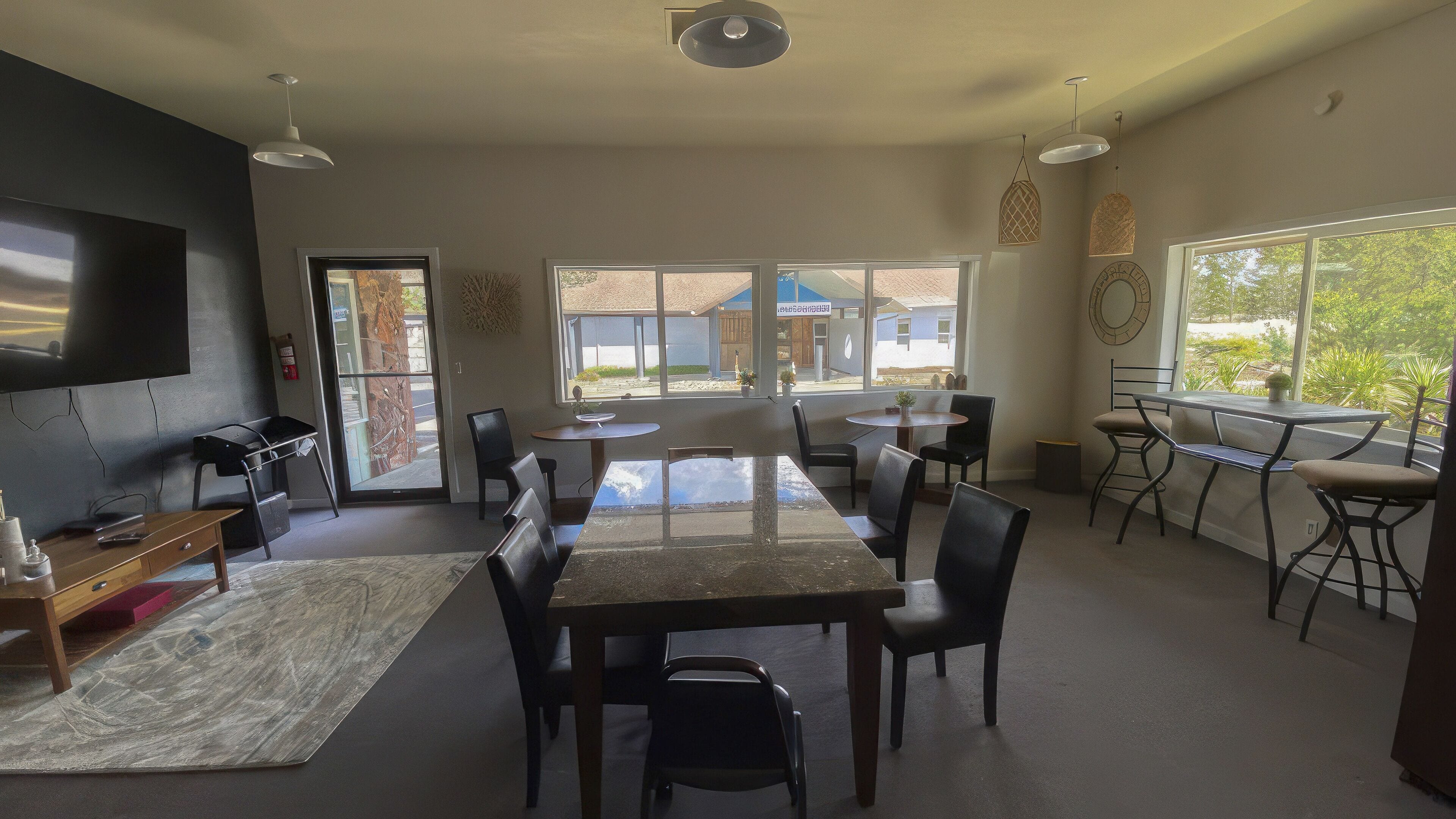 A spacious room with a large table and chairs, a flat-screen TV mounted on the wall, and large windows offering a view of the outside.