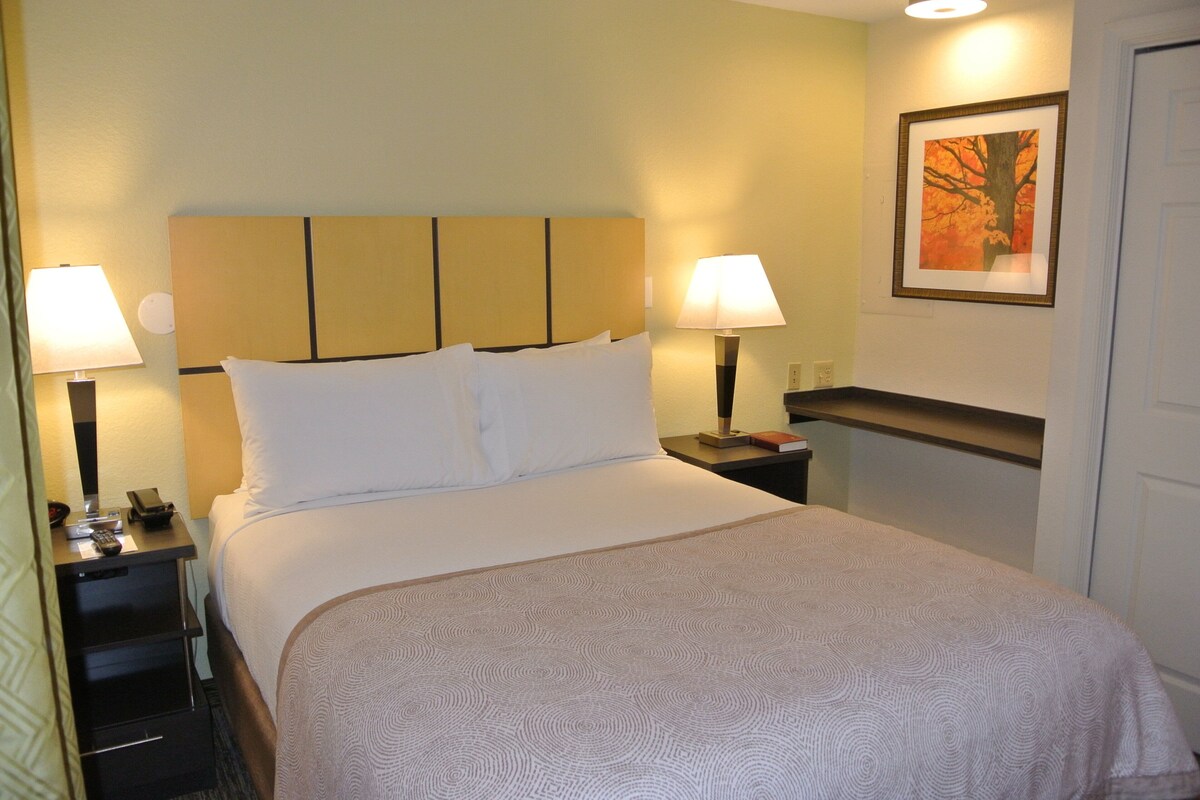 A neatly made bed with white linens, two bedside lamps, a nightstand with a phone and a book, and a framed picture on the wall.