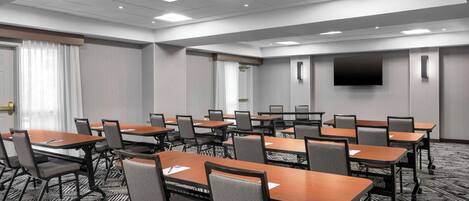 A conference room with multiple tables and chairs, a flat-screen TV on the wall, and a carpeted floor.