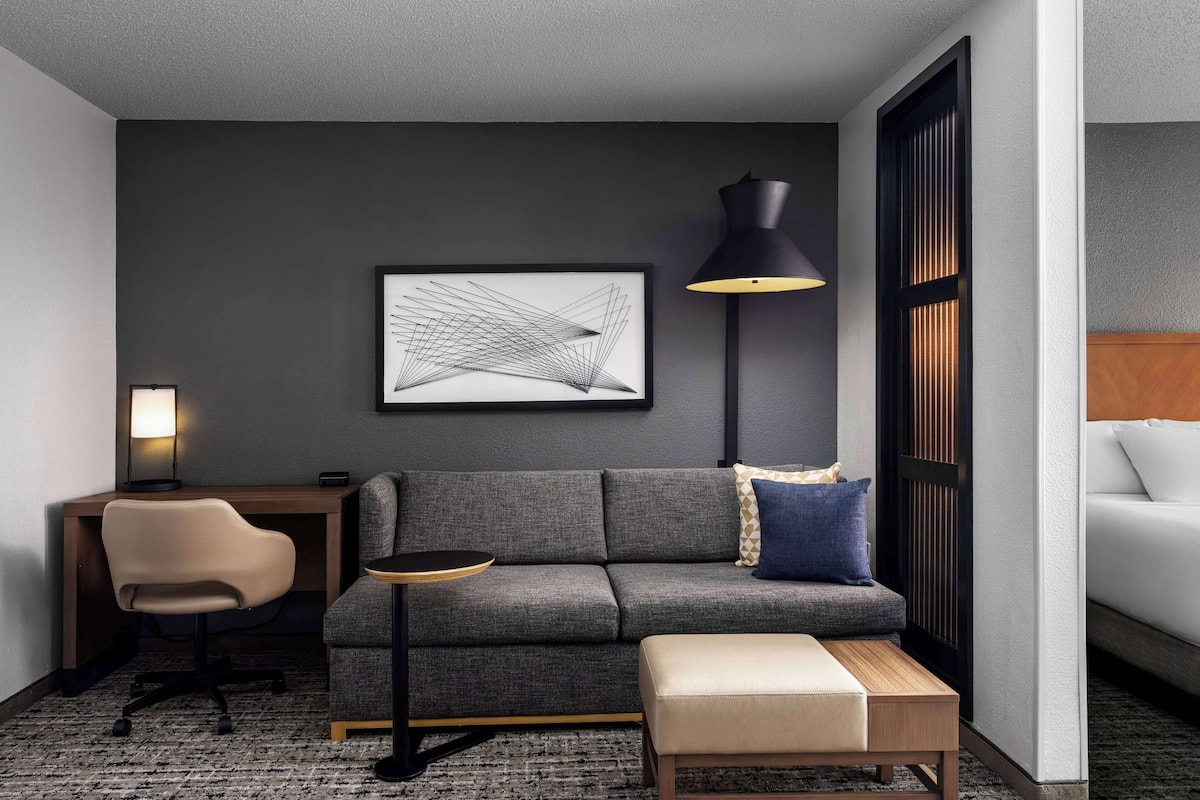 Une chambre d’hôtel moderne avec un canapé gris, une petite table ronde, un bureau avec une chaise, une lampe sur pied et une grande œuvre d’art abstraite accrochée au mur.