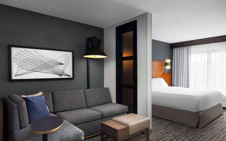 A hotel room with a gray sofa, a small wooden table, a bed with white linens, a black floor lamp, and a framed abstract artwork on the wall.