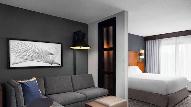 Een hotelkamer met een grijze bank, een klein houten tafeltje, een bed met witte lakens, een zwarte vloerlamp en een lijst met abstracte kunst aan de muur.