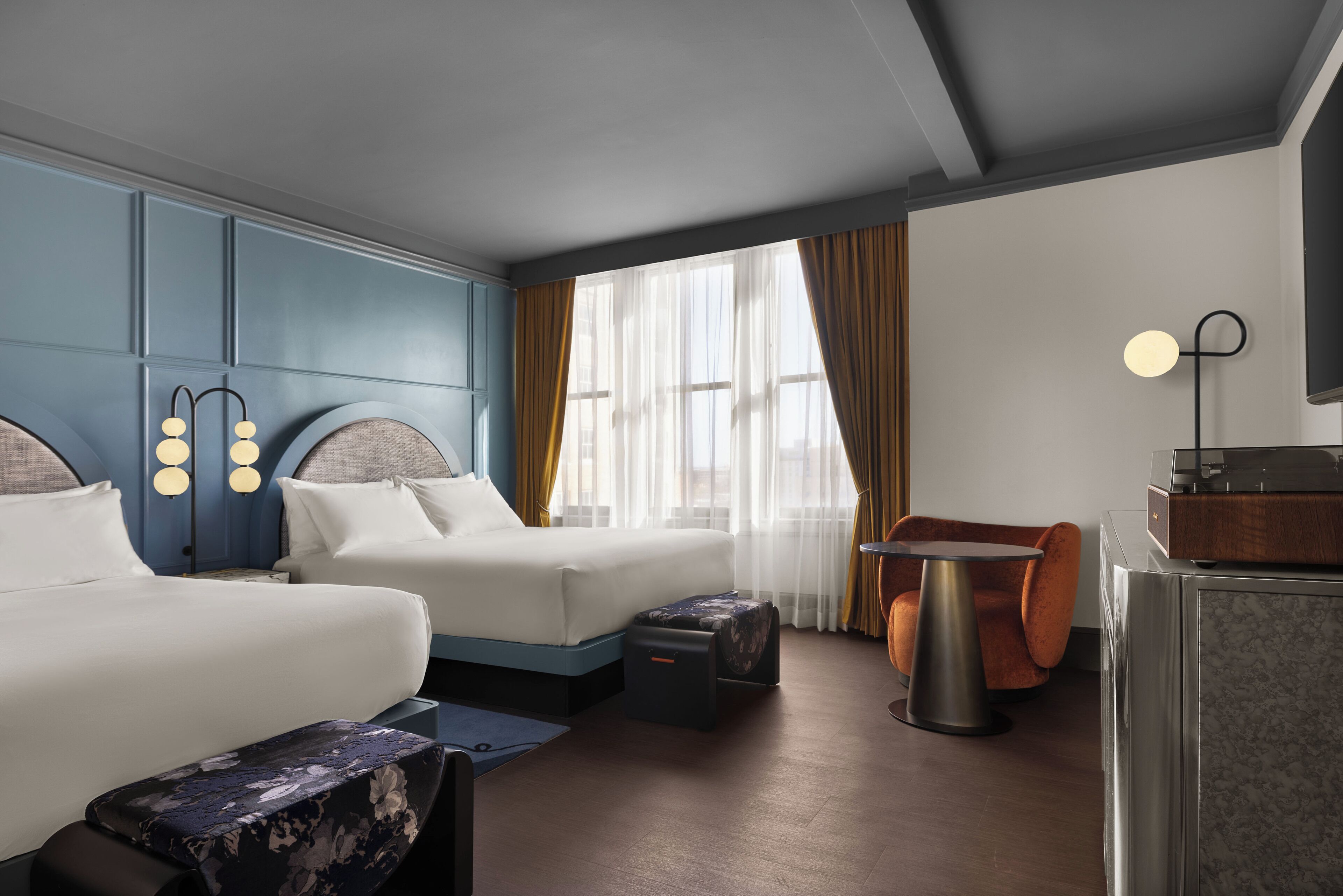 A hotel room with two beds, a small table, a chair, and a desk with a radio.
