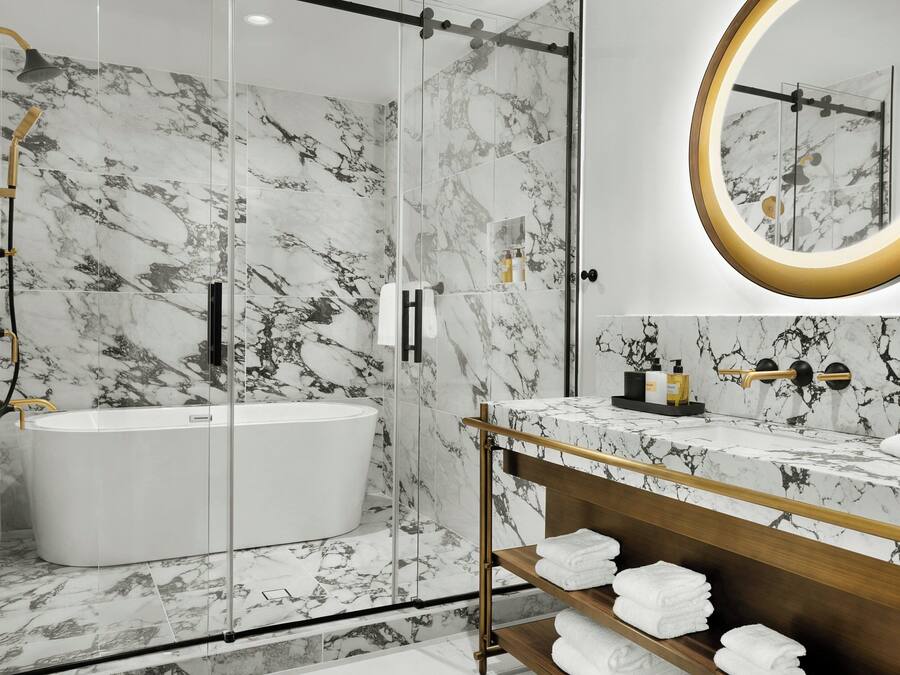A modern bathroom with a glass-enclosed shower, a freestanding bathtub, marble walls, and a vanity with a gold-framed mirror.