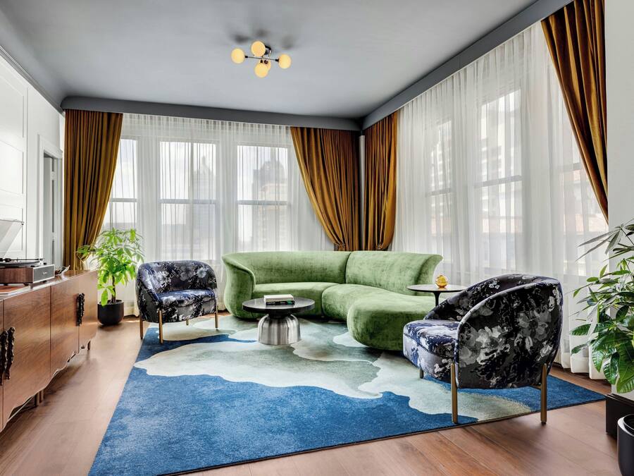 En moderne stue med en grøn hjørnesofa, to mønstrede lænestole, et sofabord, et træskab og et stort vindue med lette gardiner.