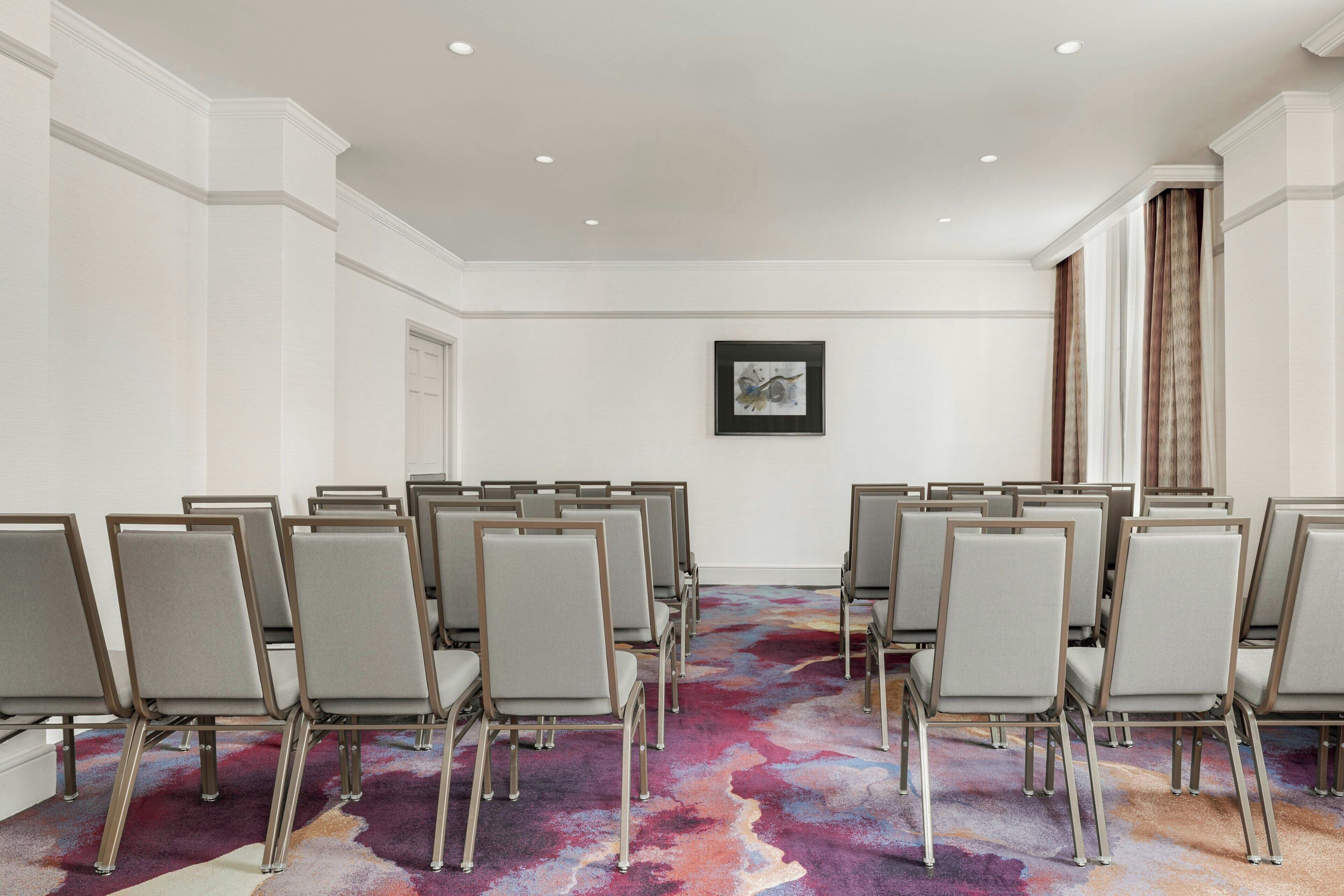 A conference room with rows of chairs, a colorful carpet, white walls, and a framed picture on the wall.