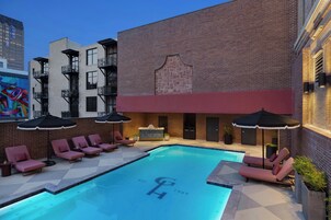 Un área de piscina en la azotea con sillones reclinables y sombrillas, un edificio de ladrillo con un logotipo distintivo y vistas a un paisaje urbano.
