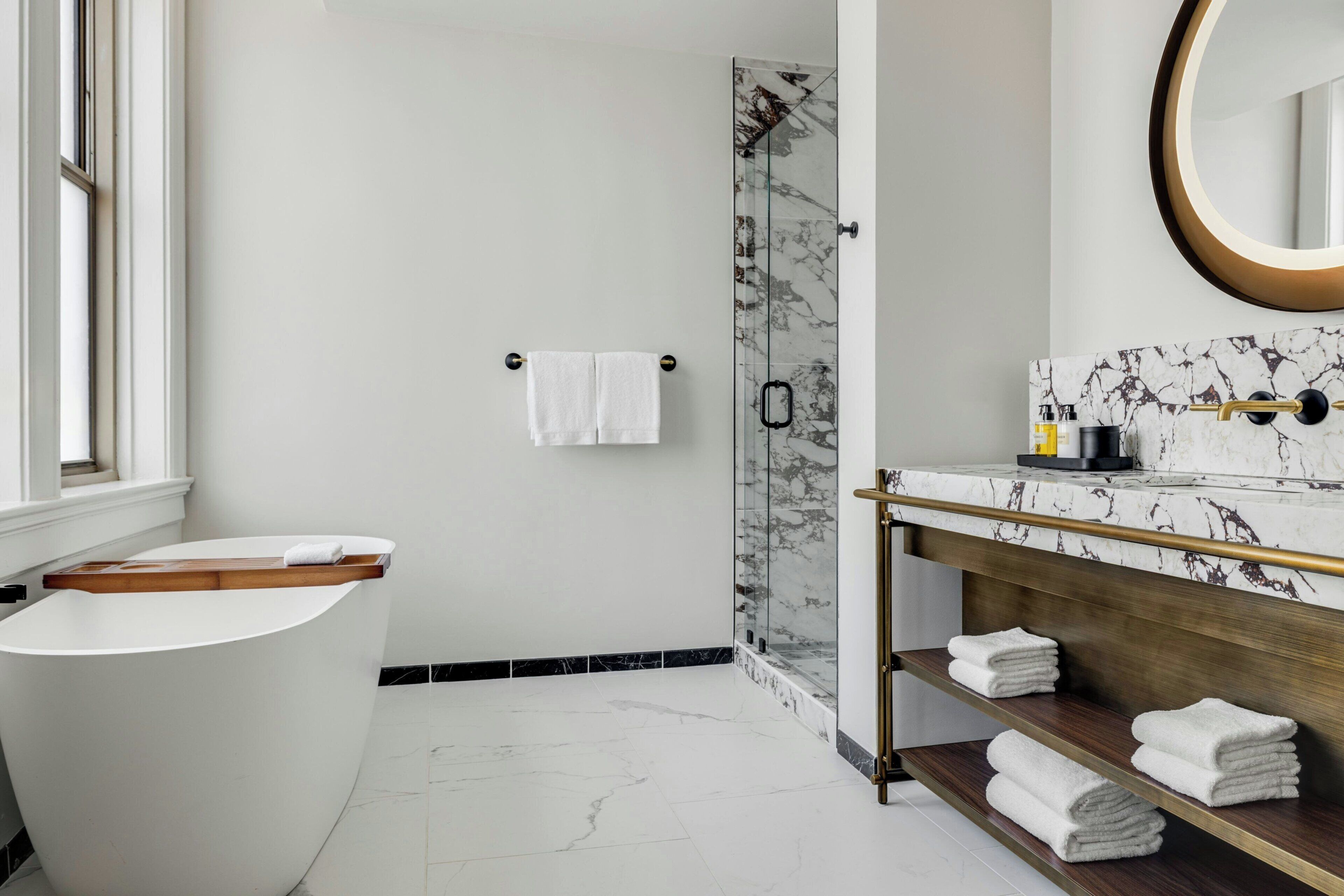 A modern bathroom with a freestanding bathtub, a glass shower enclosure, a marble-tiled vanity, and a wooden shelf with towels.