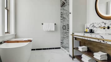 A modern bathroom with a freestanding bathtub, a glass shower enclosure, a marble-tiled vanity, and a wooden shelf with towels.