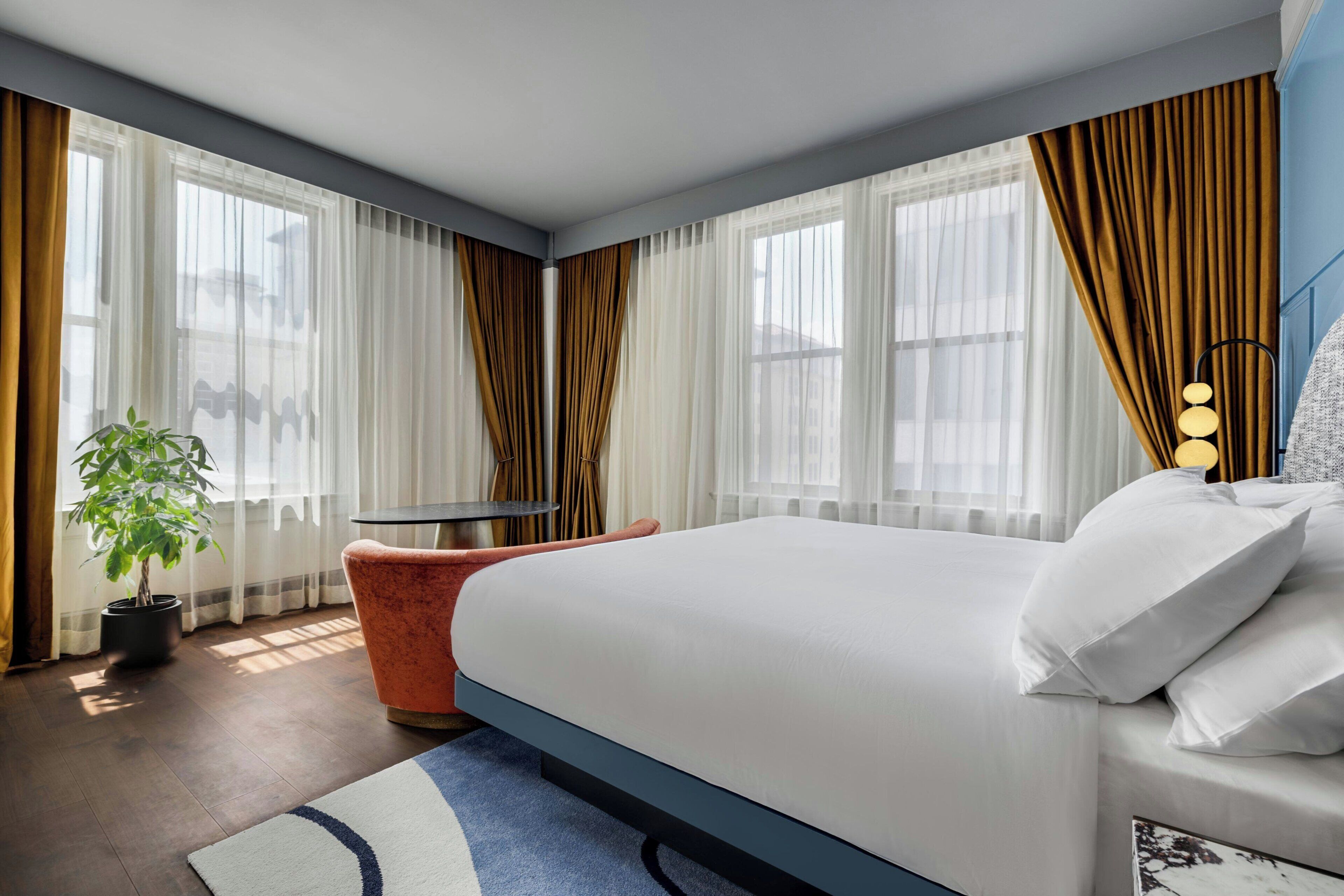 A modern hotel room with a large bed, a small round table, a potted plant, and a view of the city through large windows.