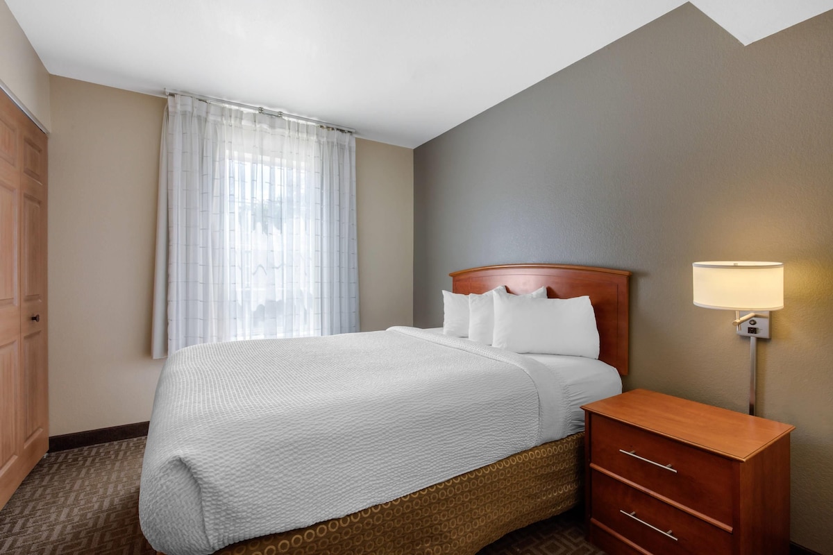 Une chambre d’hôtel avec un lit, une table de chevet, une lampe et une fenêtre avec des rideaux légers.