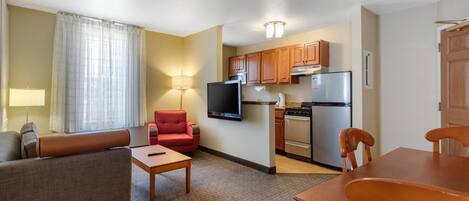 A compact hotel room with a kitchenette, a sofa, a chair, a small table, and a TV.