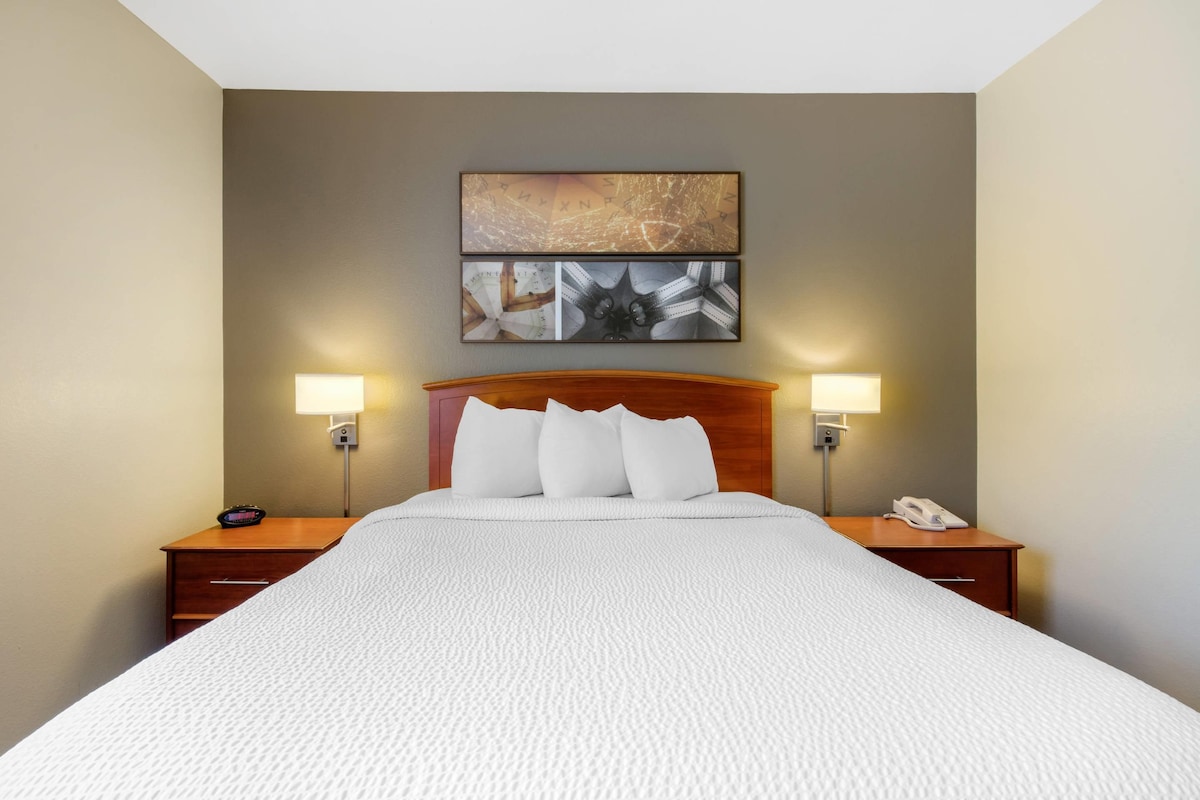 Une chambre d’hôtel avec un grand lit, deux lampes de chevet, une tête de lit en bois et une œuvre d’art accrochée au mur au-dessus du lit.