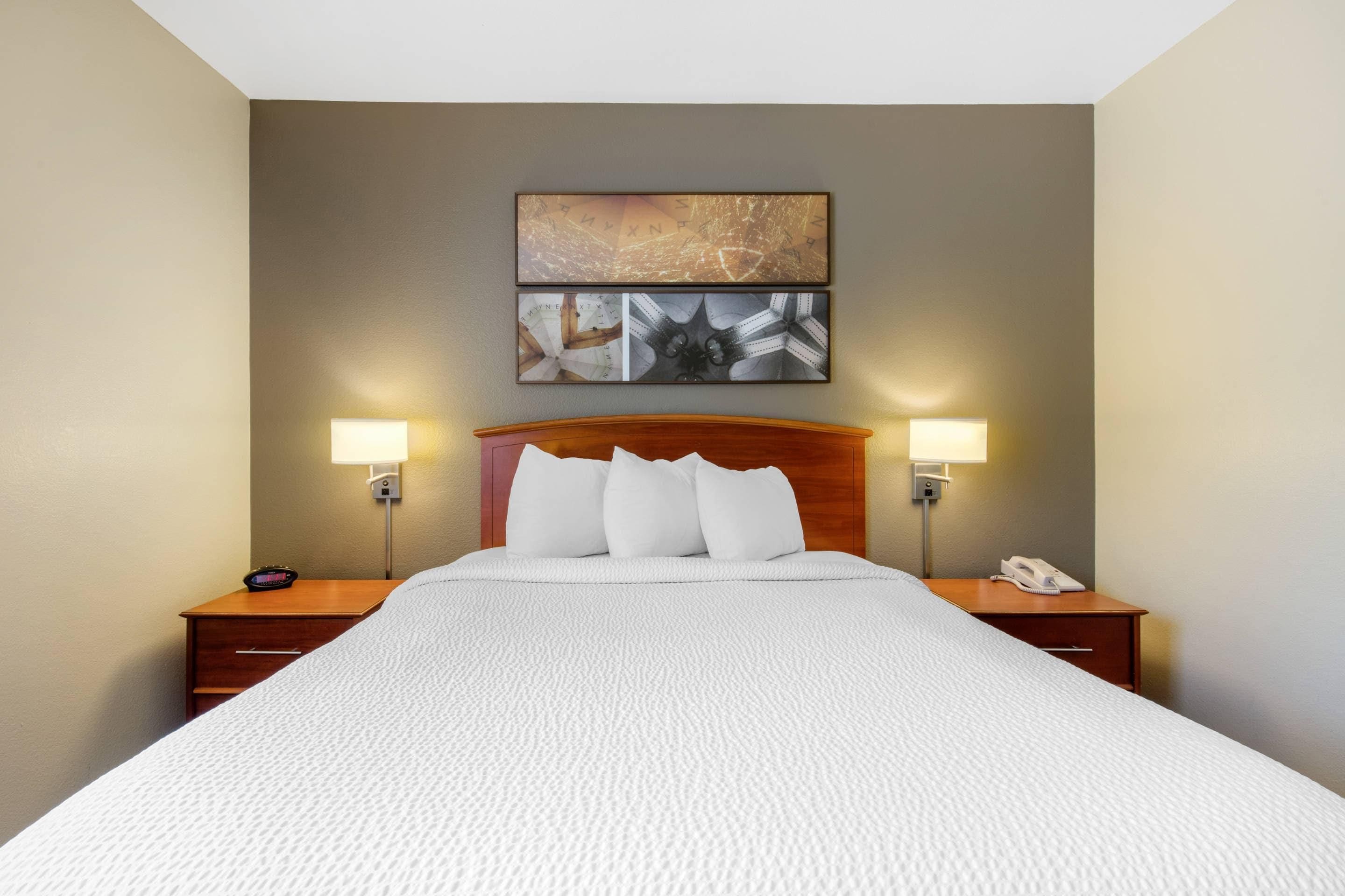 A hotel room with a large bed, two bedside lamps, a nightstand with a phone, and a wall-mounted artwork above the bed.
