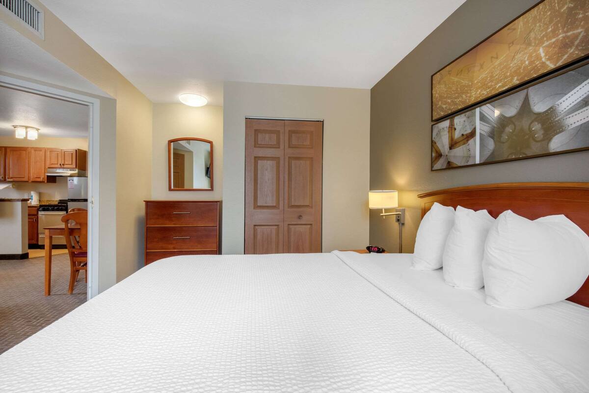 Ein Hotelzimmer mit einem großen Bett, einem Holzkopfende, einem Spiegel, einer Tür und einem durch eine offene Tür einsehbarer Küchenbereich.