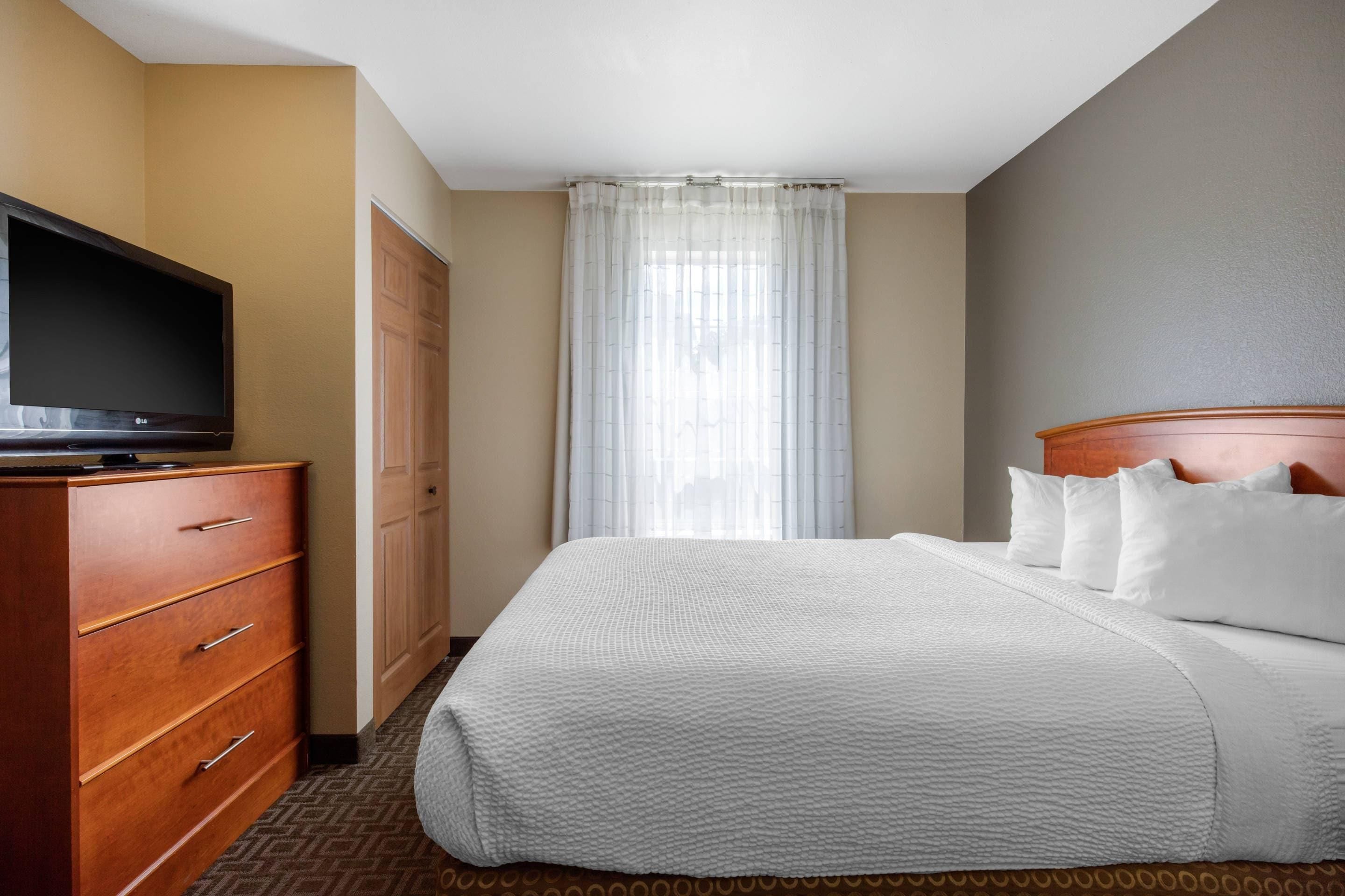 A hotel room with a bed, a television on a dresser, a wardrobe, and a window with curtains.