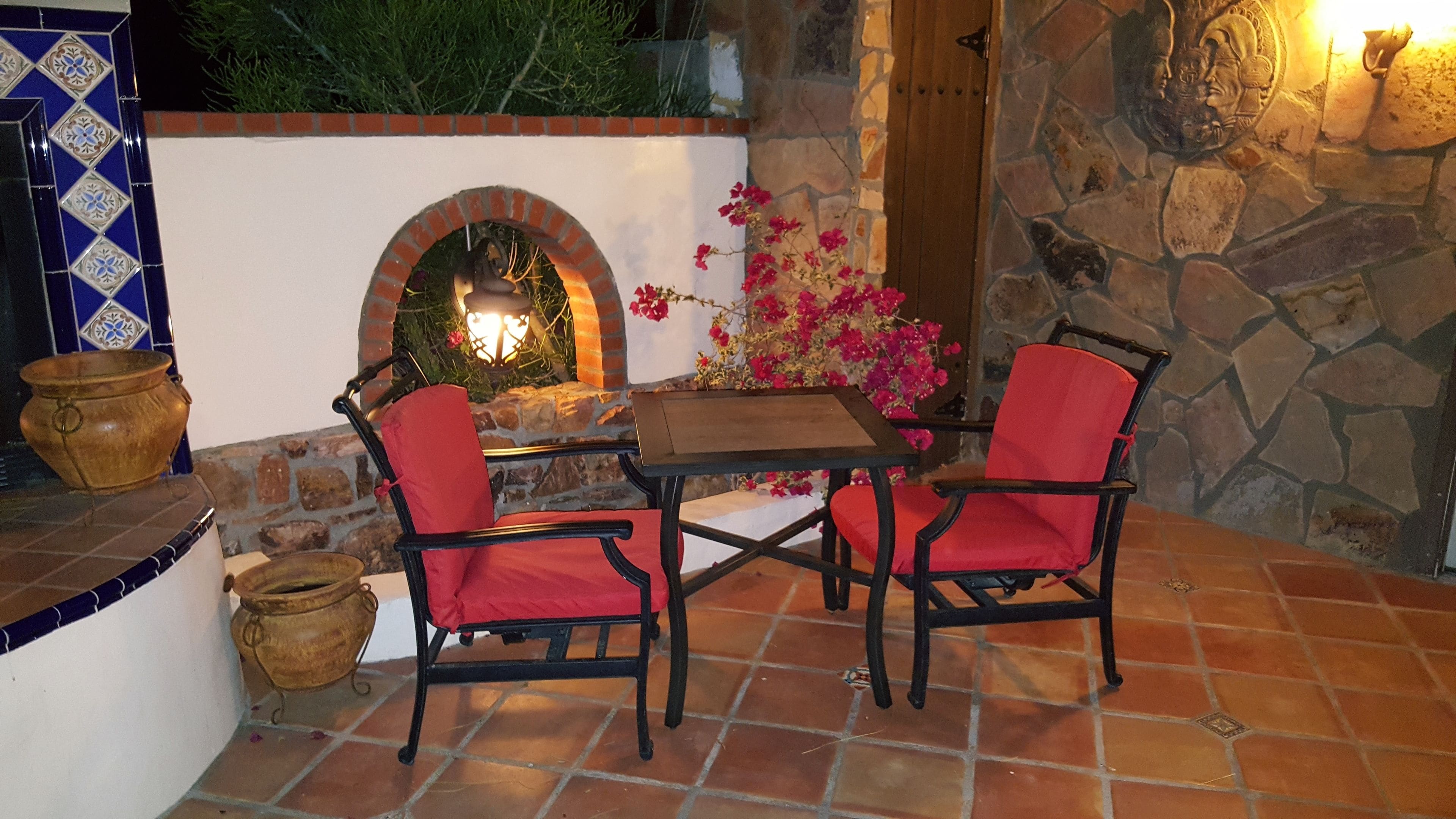 An outdoor patio with a brick fireplace, two red cushioned chairs, a wooden table, and decorative pots.