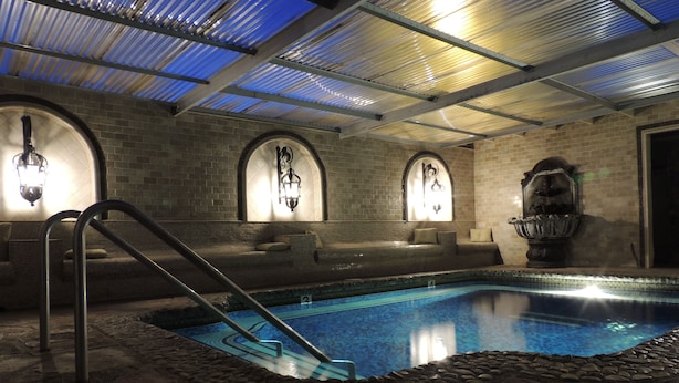 Una piscina interior con borde de piedra, escalera metálica y pared de ladrillo con ventanas de arco y luces empotradas.