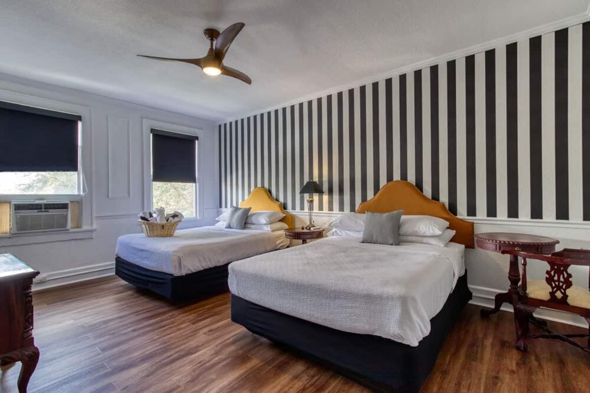 A hotel room with two beds, a ceiling fan, striped wallpaper, and a window with blackout curtains.