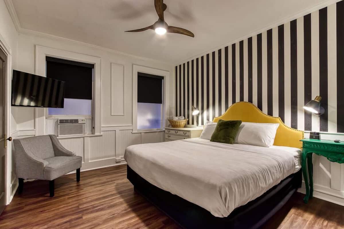 A bedroom with a bed, a chair, a ceiling fan, a TV, and a window with blinds.