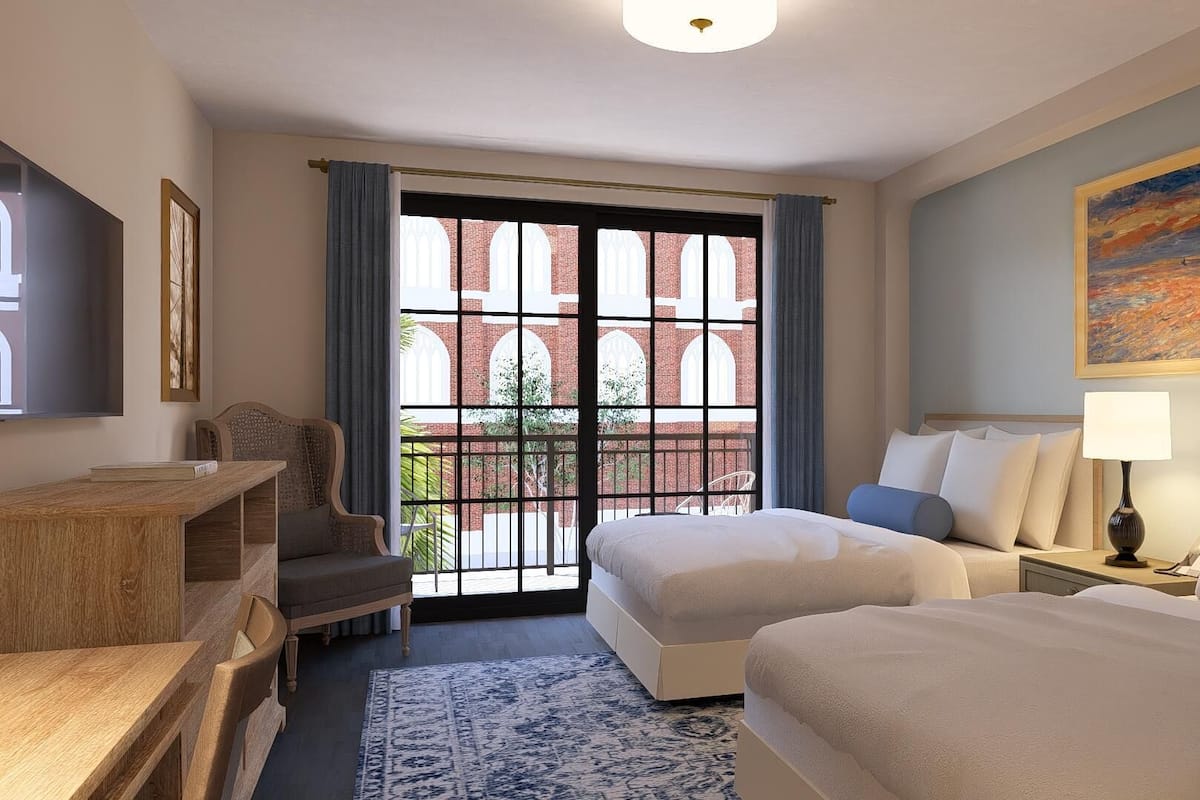A hotel room with two beds, a desk, a chair, a painting, and a view of a brick building through a window.