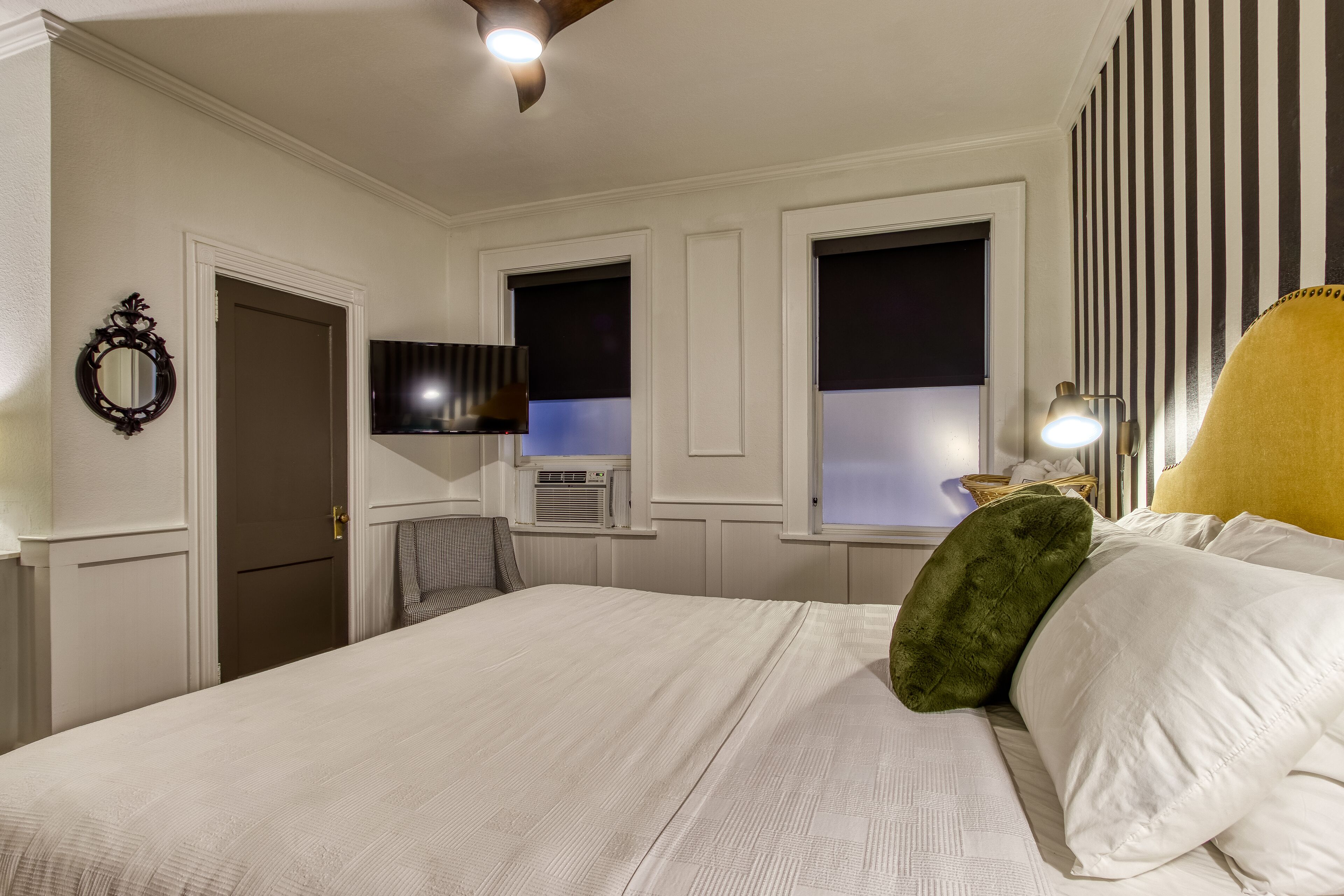 A hotel room with a large bed, a flat-screen TV mounted on the wall, a window with blackout curtains, a bedside lamp, and a mirror.