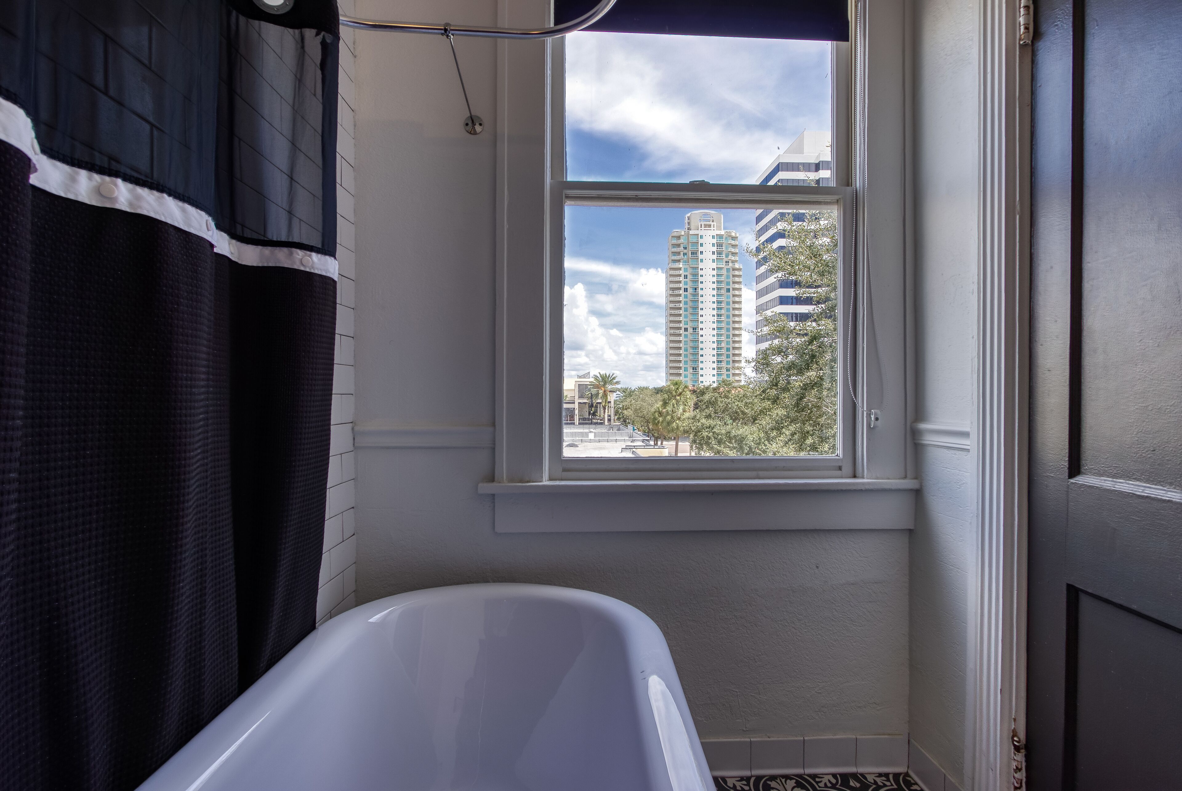 Een badkamer met een bad, een raam met uitzicht op hoge gebouwen en een deur.