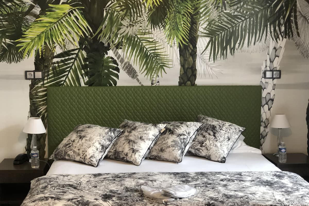 A bed with a floral bedspread and matching pillows, a green quilted headboard, and a wall with tropical plant decorations.