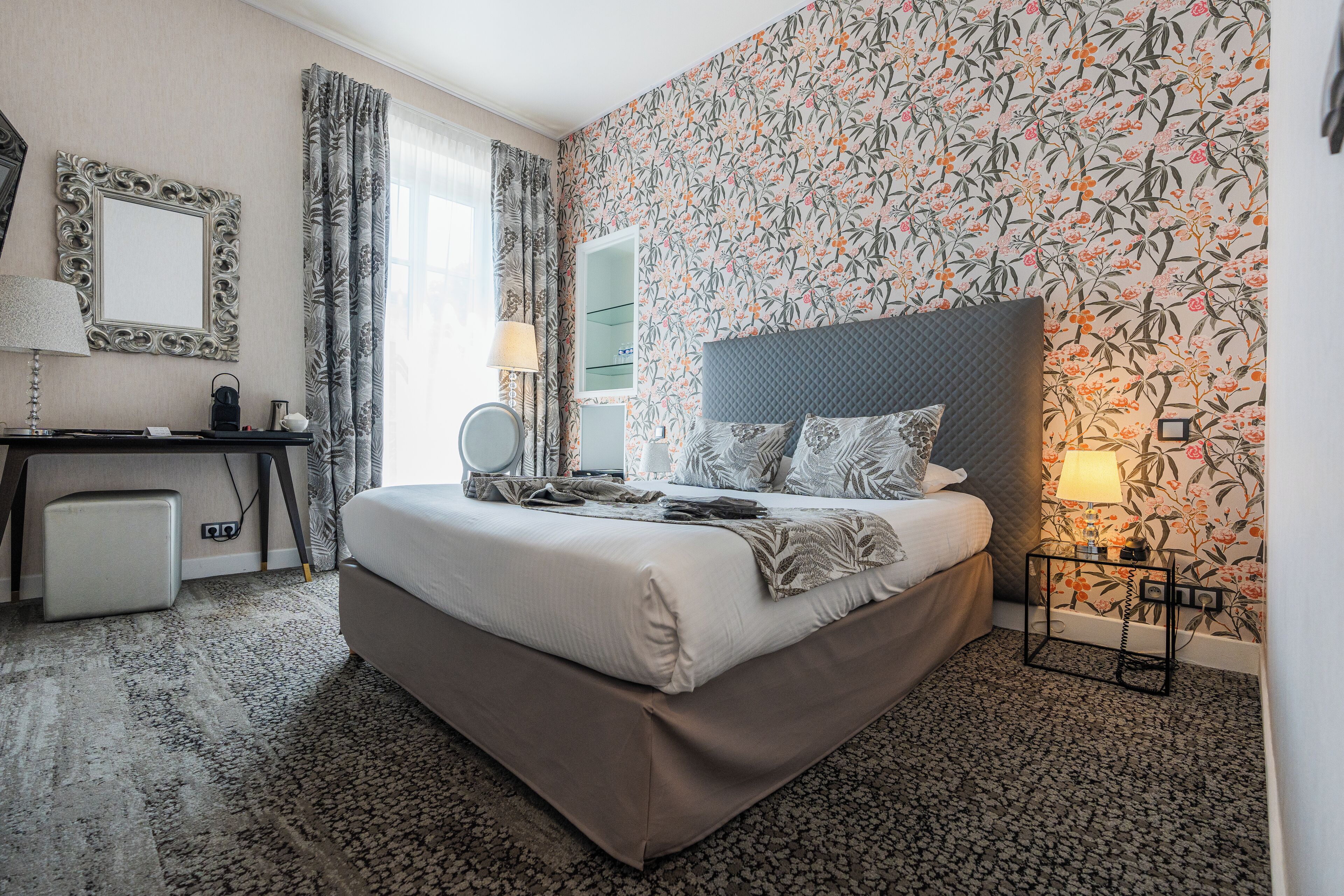 Quarto de hotel com uma cama grande, uma escrivaninha com abajur, um espelho e papel de parede floral.