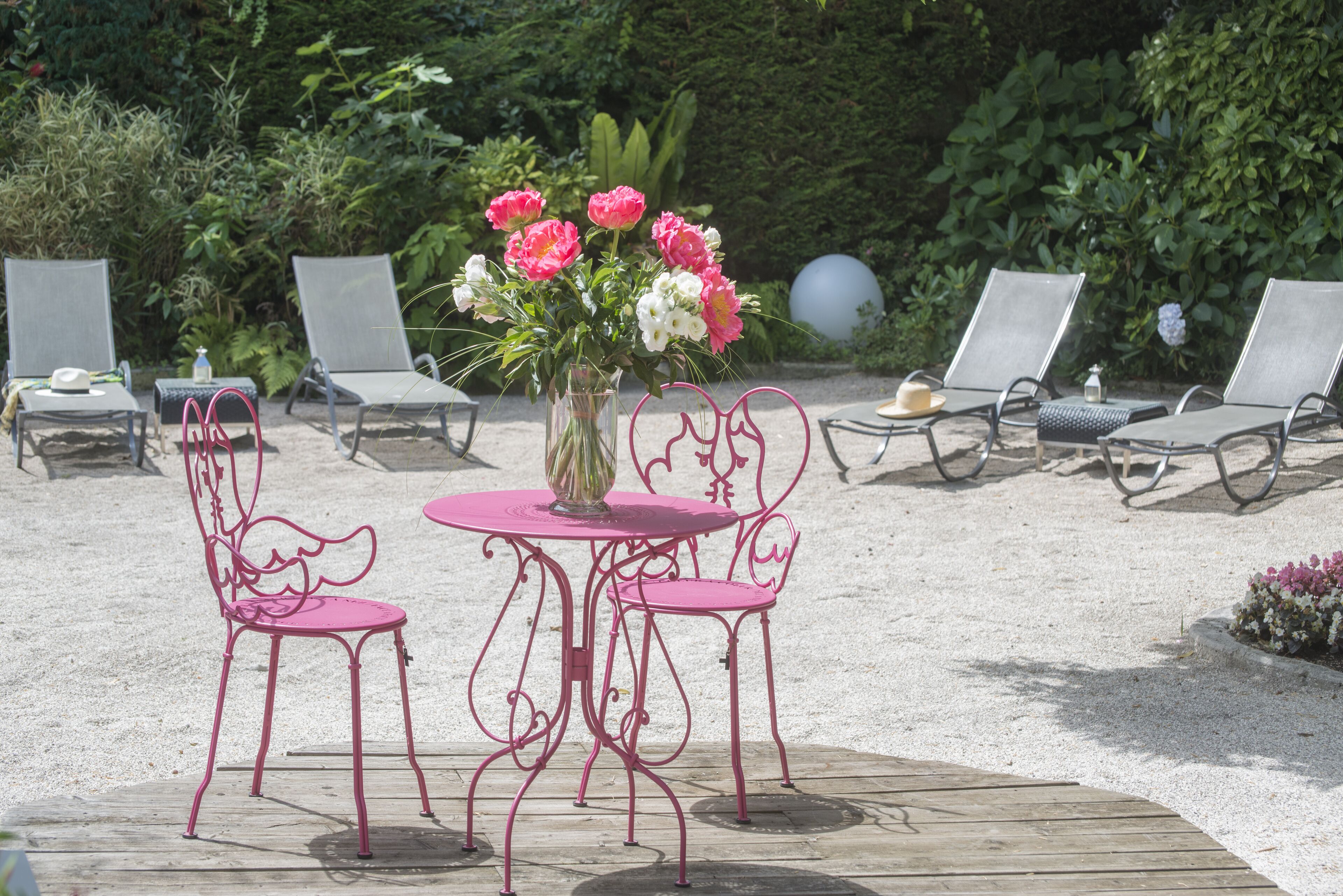 Um pátio com duas cadeiras de metal cor-de-rosa e uma mesa, cercado por vegetação e um vaso decorativo com flores.