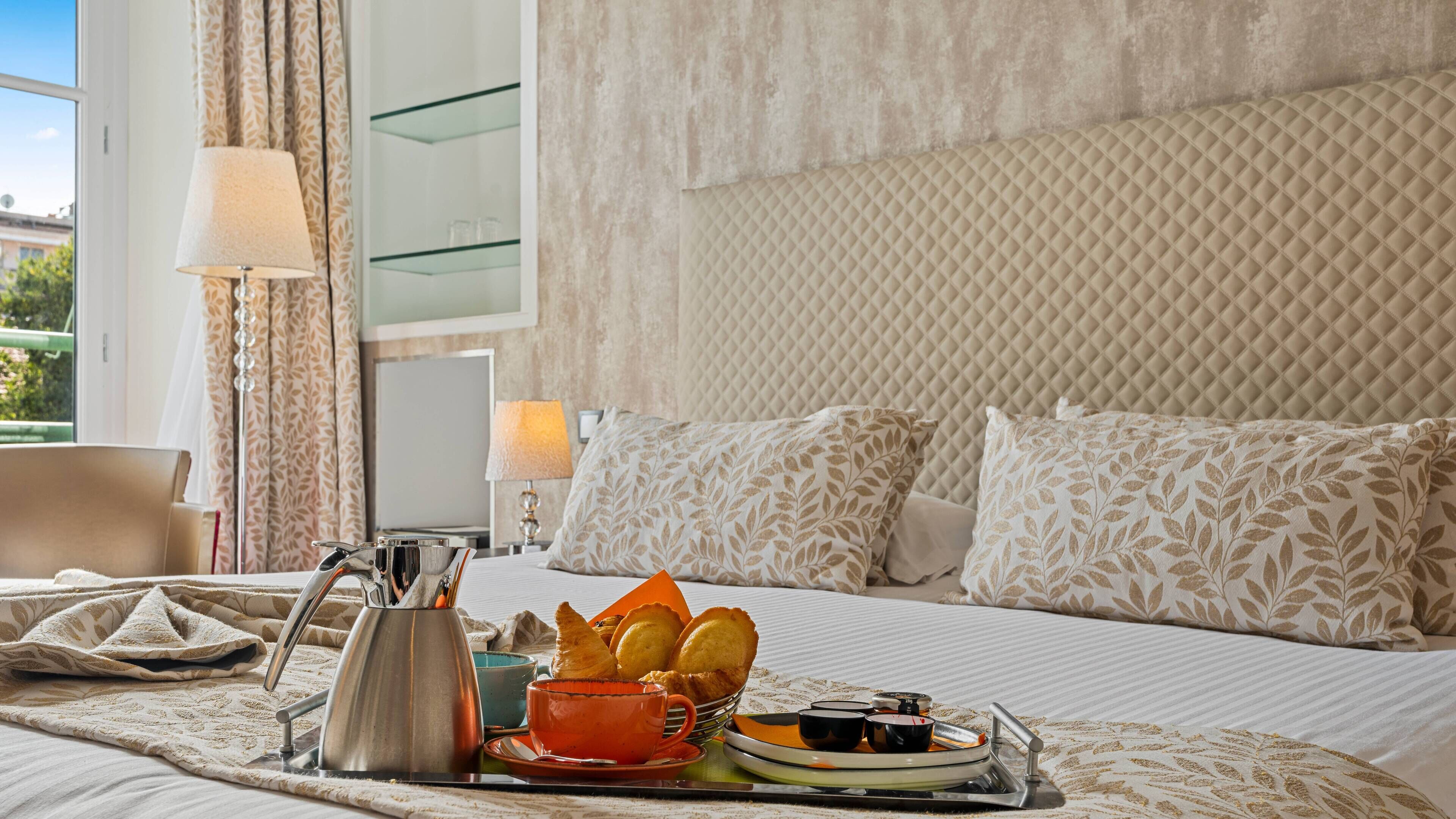 Habitación de hotel con cama, mesita de noche con lámpara, bandeja con alimentos para el desayuno y vista al exterior.