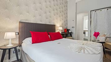 Ein Hotelzimmer mit einem großen Bett, zwei roten Kissen, einem Nachttisch mit Lampe sowie einer Kaffeemaschine auf dem Nachttisch und einem durchgegangenen Türspalt sichtbaren Badezimmer.