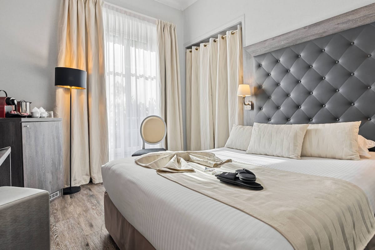 A neatly made bed with a tufted headboard, a bedside table with a lamp, and a window with sheer curtains.