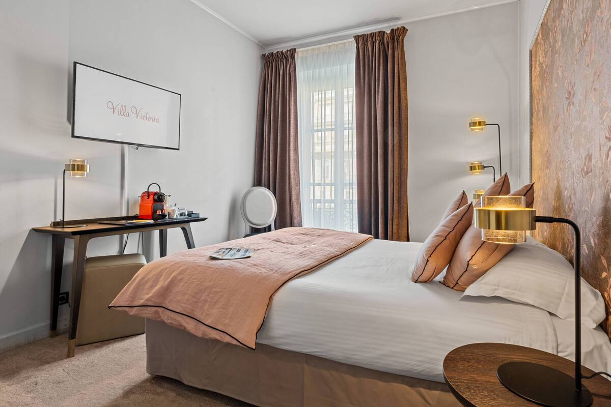 A hotel room with a bed, a desk, a chair, a bedside table, a lamp, and a wall-mounted TV.